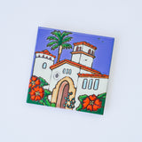 The Courthouse of Santa Barbara Art Trivet is a hand-painted ceramic tile showcasing a stylized white building with red-tiled roofs, arched doorway, palm tree, and bright red flowers against a blue sky, inspired by the county courthouse.