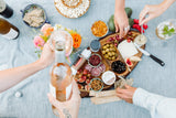 A picnic scene with hands reaching for food and pouring wine over cheese, olives, fruits, nuts, Crackers & Spreads, and flowers on a blue tablecloth.