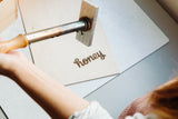 A person uses the Custom Logo Box Brand to burn the word "honey" in cursive onto a square piece of light-colored wood, creating a personalized logo with this custom branding iron.