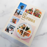 Eat California: Vibrant Recipes from the West Coast, a California cookbook, sits on a white surface. The cover shows California scenery, dishes, and food trucks in a stylish grid pattern, reflecting the essence of West Coast cuisine.