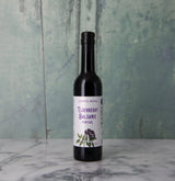 A dark glass bottle of Elderberry Balsamic Vinegar, made with upcycled elderberries and featuring a white label with purple details, stands on a marble surface against a textured green background.