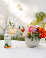 A glass of iced elderflower drink with mint sits beside a bottle of Elderflower Syrup on a white table with colorful flowers, set against a blurred outdoor background.