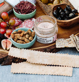A charcuterie board with Everything Spice Sourdough Flatbread Crackers, almond pâté, grapes, sliced salami, figs, seasoned almonds, olives, seeded crisps, and a bowl of tapenade beautifully arranged on a wooden board.