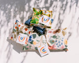Four small Eye Pillows in Strawberry Fields Canvas, each with vibrant floral patterns and labeled “Insert Thread” and “bespoke sachet buds,” are arranged on a light surface with leafy shadows.