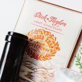 Close-up of a Dick Taylor Ginger Snap Milk Chocolate bar, a bean-to-bar seasonal release inspired by ginger snap cookies, partially obscured by a dark bottle in the foreground.