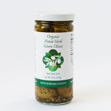 A glass jar labeled Herb Green Olives with green and white graphics, featuring whole Santa Barbara olives, California grown herbs, and a net weight of 8 oz. The label highlights its Santa Barbara County origin.