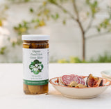 A jar of Herb Green Olives, infused with California-grown herbs, sits on a white table beside a plate of sliced salami, figs, and olives. The softly blurred background features greenery and sunlight.