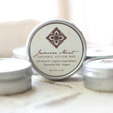 A round metal tin labeled Jasmine Mint Organic Lotion Bar sits on a light wooden surface. This all-natural, vegan massage bar is handmade with organic ingredients, jasmine floral wax, and mint essential oil.