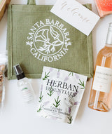 A gift set includes a green Santa Barbara, California tote bag, a welcome card, a bottle of rosé wine, Lavender Essential Oil Towelettes, a spray bottle, and a tube of nuts—making it the perfect travel kit.