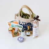 The Local Flavors Santa Barbara Traveler Tote is a travel-ready gift set with a jute bag, postcards, chocolate truffles, jam, spray bottle, olive oil, bath salts, and more—all artfully displayed on a white background.