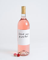 A bottle of Love You Bunches Rosé Natural Wine with a minimalist white label and cork rests on a white surface next to two small pink flowers.