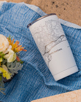 The Map of Santa Barbara Insulated Tumbler with a white body and black engraved design sits on a blue textured cloth near flowers and a sandy surface.