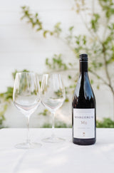 A bottle of Margerum Santa Barbara County M5 Rhone Blend sits beside two empty glasses on a white tablecloth, with a soft-focus background of greenery and a white wall.