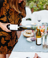 A woman in a patterned black and orange dress pours Margerum Santa Barbara County M5 Rhone Blend into a glass at an outdoor table set with wine bottles, flowers, and snacks.