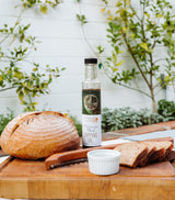 A loaf of rustic bread, sliced with a wooden-handled knife, rests on a board next to a small white dish and Mediterranean Herb Dipping Oil. Lush green plants and a white fence make the scene inviting.