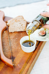A hand pours Mediterranean Herb Dipping Oil into a ramekin on a wooden cutting board, surrounded by bread slices, fresh basil, a halved fig on a plate, and a bread knife.