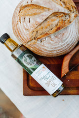 A round loaf of rustic bread rests on a wooden board beside a bottle of Mediterranean Herb Dipping Oil, infused with basil for a fresh, vibrant flavor.