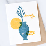 The Mediterranean Sun Thank You Letterpress Card shows a blue vase with a leafy branch, a yellow sun-like circle, and wavy yellow lines. "Thank you" in yellow script sits at the top right. Card comes with a kraft envelope.