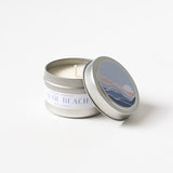 A silver tin labeled “Miramar Beach Travel Candle” sits with its lid off, showing an unlit wick. The lid displays a muted beach scene with waves, hills, and a moon, evoking the fresh scent of an ocean breeze.