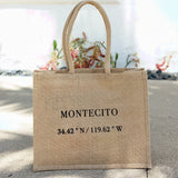 The Montecito Coordinates Market Tote, an eco-friendly beige jute bag with "MONTECITO" and 34.42° N / 119.62° W coordinates, rests on a sunlit sidewalk with plants and a white wall in the background.