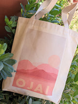 The Ojai Pink Moment Canvas Tote, crafted in the USA from organic cotton, showcases a pink mountain and sun graphic with "OJAI" below—all set against beige fabric and displayed among green leafy plants.