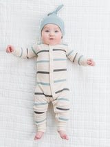 A baby in a cream striped onesie wears the Organic Knotted Baby Hat in Tide (6-12 months), the light blue hat framing their face as they look up with arms outstretched and a gentle smile on a white quilted surface.