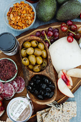 A charcuterie board with pepper stuffed green olives, black olives, salami, cheese, red grapes, apple slices, crackers, almond pâté, olive tapenade from Santa Barbara County olives, avocados, and an 8 oz bowl of granola on a wooden board.