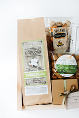 A gift basket with Petite Sea Salt & Olive Oil Santa Barbara Popcorn, gluten-free organic pistachios, California-crafted herb almonds, and a small brown gift box tied with a green ribbon.