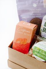 A close-up of the Petite Sweet & Savory Box featuring a pink box of California Gummy Bears, dark chocolate granola cookies, and California almonds in vibrant packaging—all artisan snacks arranged beautifully.