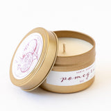 The Pomegranate Travel Candle is a gold tin with a pomegranate-illustrated lid set aside, revealing white soy wax inside. The label reads "pomegranate fragrance" and "3 oz." Compact and ideal for travel.