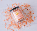 A glass jar of Poppy Bath Salts, with a black lid and a peach floral label, lies on its side as pink Himalayan bath salts infused with citrus and geranium oils spill onto a white surface.