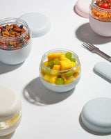Three small, round Porter Seal Tight Bowls in Mint, filled with snacks—one with mixed nuts, one with fresh fruit, and one partially visible—sit on a white surface with matching pastel lids and a silver fork.