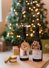 Two bottles of Rangeland Mistletoe Red Blend with festive tags sit on a table beside a glowing candle, with a softly lit Christmas tree in the background—an ideal holiday wine gift scene.
