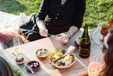 At a picnic, someone spreads dip on bread with Riverbench Blanc de Blanc Sparkling Wine and snacks outdoors.