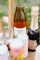A close-up of Riverbench Brut Rosé, a blush pink sparkling wine, with a wine glass and colorful cans in the foreground, set outdoors.