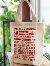 The SB Local Canvas Tote is a natural cotton bag with sturdy handles, featuring vibrant red text in various fonts naming local spots like Gaviota and Lotus Land. Shown here resting on a wooden surface.