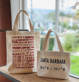 Two SB Local Canvas Tote bags sit by a window. The left features red text listing local spots; the right shows “Santa Barbara” with coordinates in black. Both have a local destinations print, are made in USA, with houses and greenery outside.
