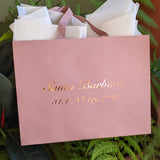 The Santa Barbara Coordinates Blush Pink Gift Tote, featuring “34.4°N 119.7°W” in gold, sits among green ferns with white tissue peeking out—a stylish, eco-friendly choice for a wedding welcome gift.