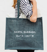 A person in a black and white striped shirt holds the Santa Barbara Coordinates Jute Tote - Blue, featuring "SANTA BARBARA 34.4° N / 119.7° W" in white letters on the front.
