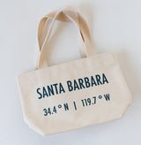 The Santa Barbara Coordinates Petite Canvas Tote is a beige canvas bag with short handles, made in the USA. It features "SANTA BARBARA" and 34.4° N | 119.7° W in bold black letters and is displayed on a white surface.