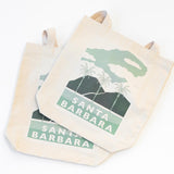 Two Santa Barbara Palms Canvas Totes with green and white palm tree, mountain, and cloud graphics plus "Santa Barbara" text are shown overlapping on a white background—ideal as a city tote or beach bag.