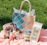 The Santa Barbara Snacks & Treats Tote rests on a pink blanket with grass in the background, featuring organic snacks such as pistachios, popcorn, wild berry mix, almond toffee, cookies, and a candle.