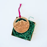 The Santa Barbara Wine Country Wood Ornament—round and engraved with a vineyard scene and “WINE COUNTRY SANTA BARBARA, CA”—comes boxed with green paper grass and a pink ribbon, making it an ideal holiday keepsake.