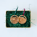 A basket with green shredded paper holds two Santa Barbara Wine Country Wood Ornaments—round wood ornaments with engraved local designs and ribbon loops—a charming holiday keepsake for any home.