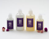 Four bottles of Santa Ynez Lavender Body Lotion, each with a purple label, are arranged in a row. Small pink and purple flowers accentuate their soothing floral scent.