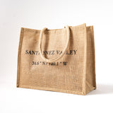 The Santa Ynez Valley Coordinates Jute Tote is a tan, eco-friendly bag with two handles, featuring "SANTA YNEZ VALLEY 34.6° N / 120.1° W" in black on the front, shown against a plain white background.
