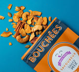 A partially opened Savoureux Snack Mix bag spills a keto blend of mixed nuts, chips, cashews, pecans, and seeds onto a bright blue surface.
