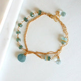 The Seaside Moss Gold Bracelet features delicate 14k gold fill, light blue moss aquamarine beads, and a teardrop pendant, shown on a white textured surface.