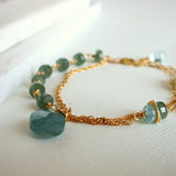The Seaside Moss Gold Bracelet features a delicate 14k gold fill chain with greenish-blue moss aquamarine beads and a faceted centerpiece, shown on a white textured surface.