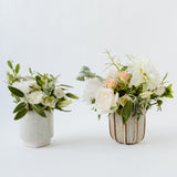 Two small ceramic vases—one speckled white, one with ribbed stripes—feature seasonal white and pale peach flowers with greenery. Perfect for local delivery: Seasonal Flowers - Cup (Local Santa Barbara Delivery Only).